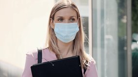 Video of a beautiful blonde woman in face mask with a folder waiting while standing in the street.