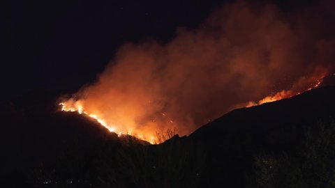 The Catalina mountains are on fire as seen in a time lapse. The fire slowly makes its way down the slope while billowing lots of smoke in 4K