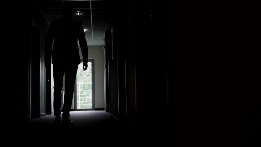 Slow motion, a backlit man walks along the corridor of an office or hotel to the glass doors, then stops and waits. Royalty-Free Stock Footage #1054661042