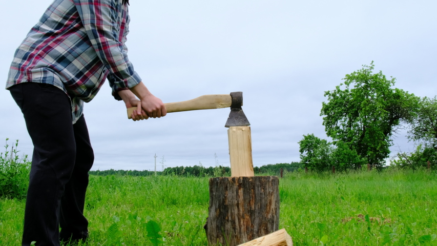 A Man in a Plaid Shirt Chops a Log with an Ax on a Grass Background.