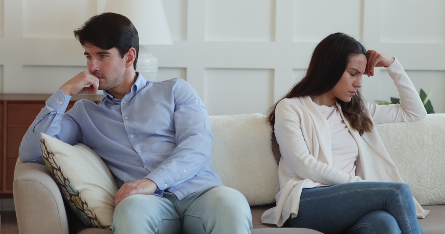 Stressed young man sitting on sofa separately from angry wife, do not talking after quarrel. Offended married couple ignoring each other, having misunderstanding indoors, family relationship problems. | Shutterstock HD Video #1054665701