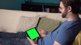 Side shot of young man with glasses waves to friends and family on tablet with green screen. 