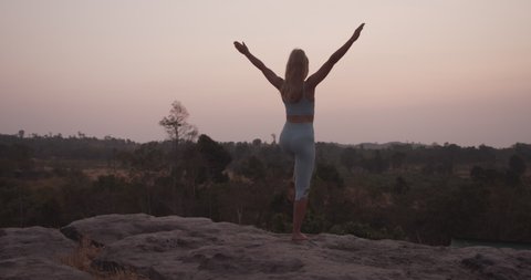 Shot of a blonde woman standing barefoot on the rocks by the mountain, with her hands raised up as she balances on one foot