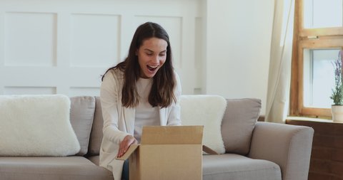 Happy positive young mixed race woman sitting on sofa, unboxing delivery carton parcel, satisfied with purchase in internet store. Smiling female client feeling excited of fast shipping service.