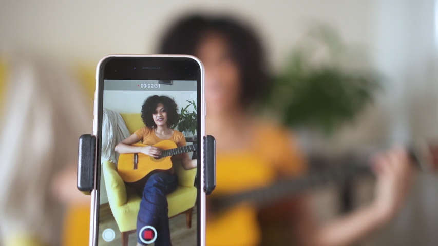 Young woman at home singing and playing guitar for followers Spbd. filming blog about learning to sing popular songs. cover, smartphone, recording. confident singer performer | Shutterstock HD Video #1054674116