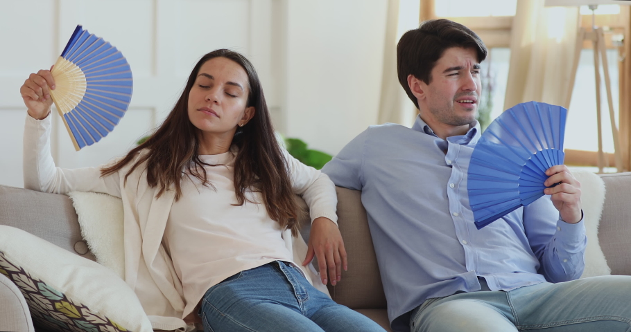 Overheated young married couple suffering from hot summer time weather or high temperature indoors without air conditioner. Displeased family spouses feeling exhausted, using paper fans at home. Royalty-Free Stock Footage #1054675181