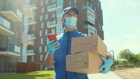 Young delivery man wear mask and gloves holding box use phone walking on street quarantine outdoor shopping package pandemic postman self isolation slow motion