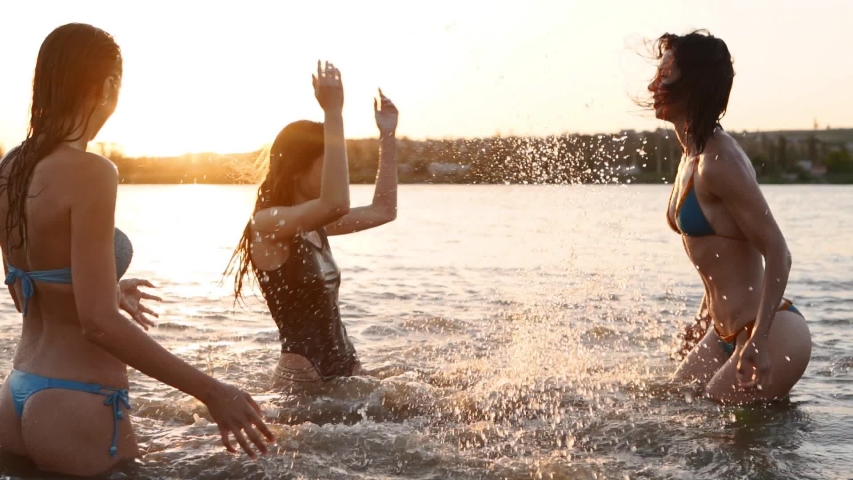 Happy wet girls in bikini run into sea and play splashing water to each other on sunset. Cheerful female friends have fun making splashes in pond. Young women go to swim, bathe in lake. Slow motion. Royalty-Free Stock Footage #1054682027