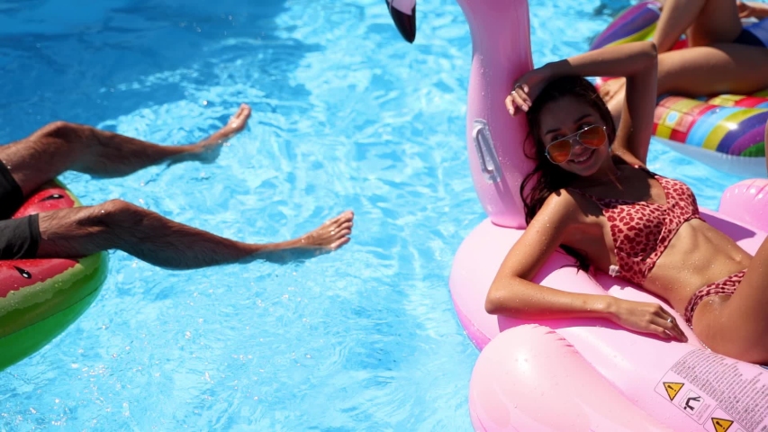 Friends chilling in private villa swimming pool, lie in the sun on inflatable flamingo, swan, floaties. Young people relax on party at luxury resort on sunny day. Bikini girls sunbathing. Slow motion. | Shutterstock HD Video #1054682045