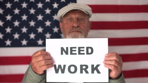 Depressed american man in a cap against the background of the USA flag holds a sign saying need work. Economic crisis, unemployment and social protests
