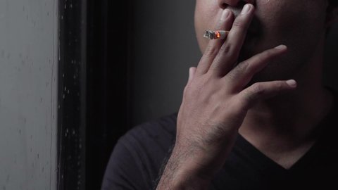 Close up handsome asian man takes a cigarette and smoking beside glass door on a rainy day. Slow motion shot