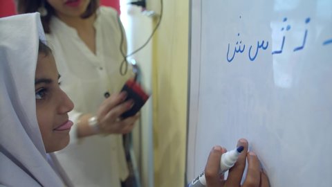 Muslim female student wearing scarf, sticks her tongue out, after making a mistake writing Urdu alphabets on the whiteboard, as teacher corrects her. Islamabad, Pakistan. 15th April 2018.