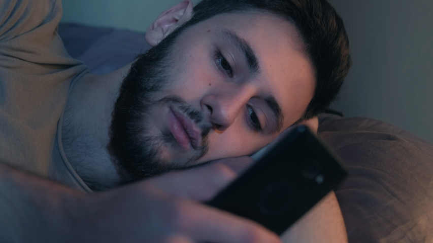 Late sleep. Night insomnia. Bored man scrolling feed on mobile phone application lying in bed. Royalty-Free Stock Footage #1054686326