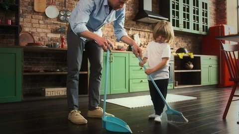 Young dad and his son in casual clothes are sweeping the floor and brushing litter into the scoop at kitchen with modern interior. Slow motion