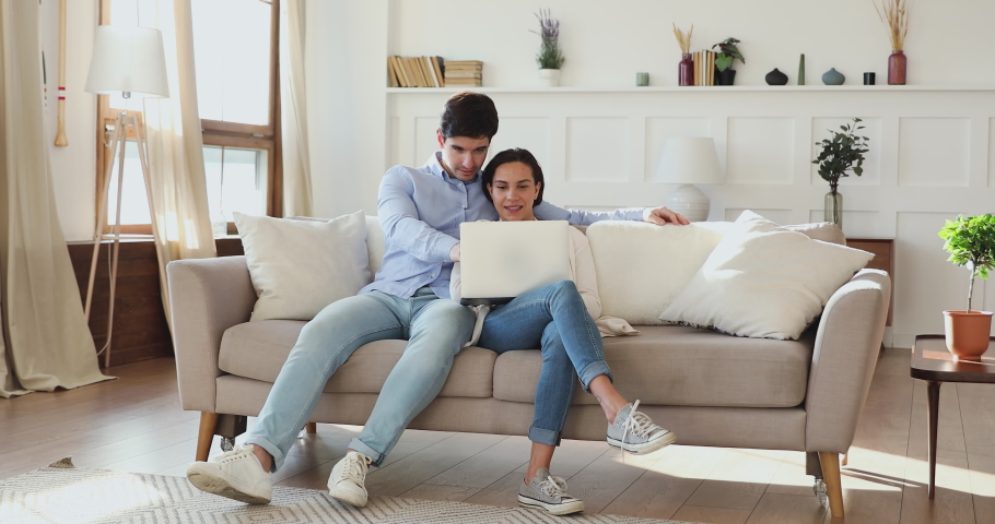 Full length affectionate loving bonding couple relaxing on sofa, looking at laptop screen, choosing summer vacation place or honeymoon trip, planning wedding or shopping online together at home. Royalty-Free Stock Footage #1054691192