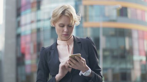 Businesswoman using her phone while standing outside office. Stylish female entrepreneur in grey suit going through her email inbox early in the morning.