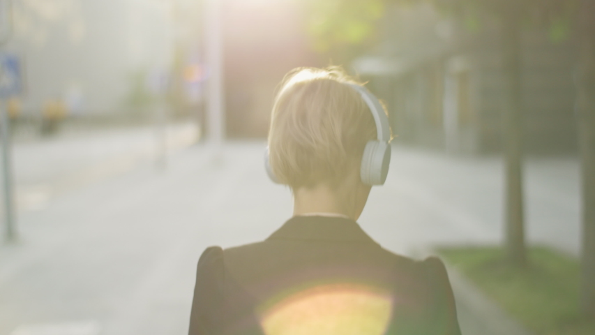 Female commuter with headphones walking on the street in the morning. Stylish woman listening to her favorite music track while getting ready for the start of a new day. Going to work. | Shutterstock HD Video #1054693142