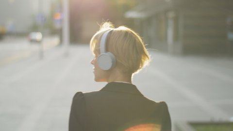 Female commuter with headphones walking on the street in the morning. Stylish woman listening to her favorite music track while getting ready for the start of a new day. Going to work.