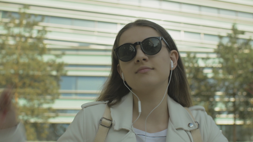 Young woman showing funny dance moves on urban street. Stylish female wearing white jacket and sunglasses listening to music outdoors. Lens flare. Royalty-Free Stock Footage #1054693589