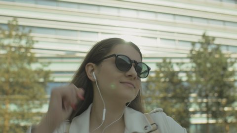 Young woman showing funny dance moves on urban street. Stylish female wearing white jacket and sunglasses listening to music outdoors. Lens flare.
