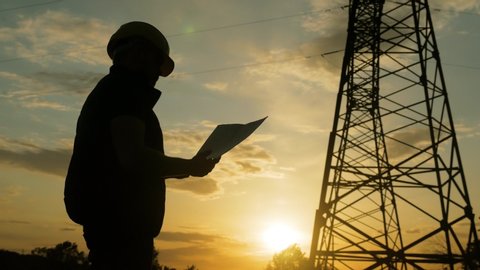 Silhouette of engineer standing on field with electricity towers. Electrical engineer with high voltage electricity pylon at sunset background. Power workers at work concept.