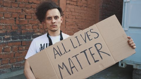 Caucasian man in mask stands against red wall with cardboard poster in hands with inscription - ALL LIVES MATTER. Single protest.
