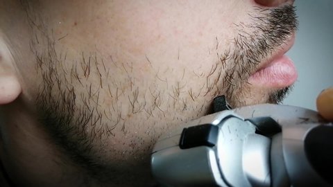 Unshaven man shaves cheek with electric safety razor. Shaving beard hairs of face. Young guy takes care of hygiene. Male hand smooth shaves stubble with shaving machine in morning close-up