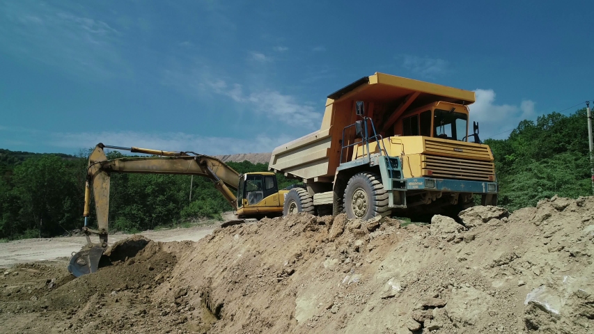 An Industrial Excavator On An Open Road Section Loads Rock And Earth On A Very Large Dump Truck, Loaded With Ore .The Excavator Bucket Drips And Loads The Ore.Panorama Of Construction Works.
 Royalty-Free Stock Footage #1054696496