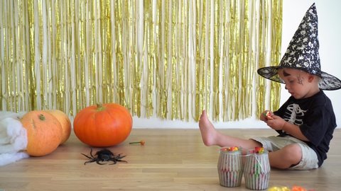 Cute 2 years old boy with magician wizard hat unpacking candy sweet sitting near pumpkins. Halloween celebration at home. Camera motion shot with gimbal.