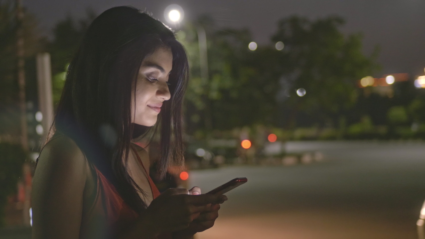 A happy and smiling corporate woman standing outside next a busy street using mobile phone to type a text message. A beautiful and attractive girl/ female freelancer on cellphone in the night