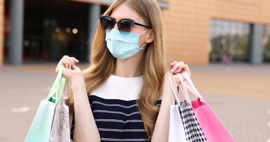 young woman in a medical protective mask on her face and sunglasses, with shopping bags, on the street in the background of a shopping center Royalty-Free Stock Footage #1054701719