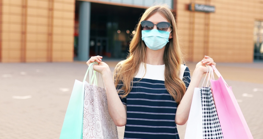  attractive woman in a medical protective mask on her face and sunglasses, with shopping bags, on the street in the background of a shopping center Royalty-Free Stock Footage #1054701734