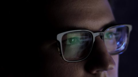 Man in eyeglasses late at night scrolling in front of laptop. Coder, programmer or developer using laptop in dark. Close up of glasses with reflection of computer screen.