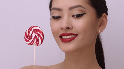 Portrait of attractive Asian woman holding colorful sugar candy, isolated on bright studio copy space background. Beauty fashion model girl with perfect makeup with big swirl lollipop in her hands