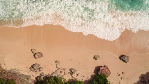 Top down aerial view from above of giant ocean waves crashing and foaming on empty sand tropical beach with big rock stones. Bird's eye aerial shot of golden beach meeting deep blue ocean water.