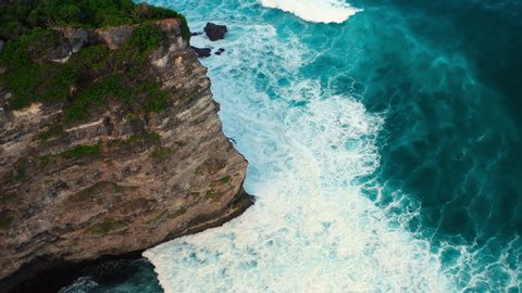 Aerial top view of waves break on rocks in a blue ocean. Sea waves on beautiful beach aerial view drone 4k shot. Bird's eye view of ocean waves crashing against an empty stone rock cliff from above.