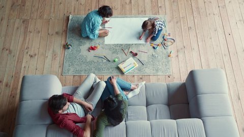 Creative activity. Top view of two little kids, brother and sister drawing with pencils, lying on the floor while young parents relaxing on the sofa