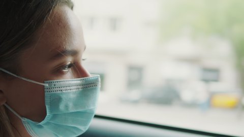 Woman wearing a medical mask in taxi car on backseat looking out of window checking cell phone. Girl passenger waiting in a traffic jam during coronavirus quarantine. Covid is over