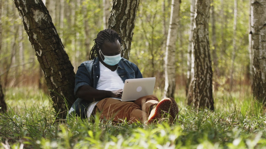 Remote work concept. African man with medical mask using laptop in the park.