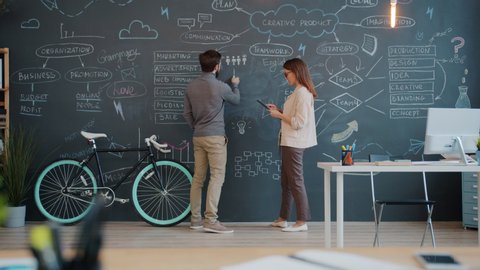 Man and woman coworkers are writing on chalk board discussing business strategy in open space office. Interior, coworking space and marketing concept.