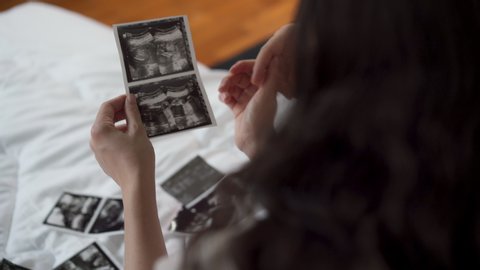 Beautiful pregnant woman holding and looking ultrasound photo of her newborn baby together with husband on the bed in bedroom. Husband and wife relationship and maternity expecting healthcare concept
