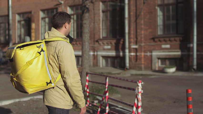 Caucasian male delivery man in a black mask and jacket with a yellow backpack and pizza boxes is standing outside in sunny weather and turns to camera and puts on a mask.
