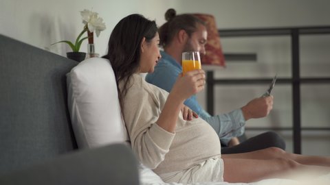 4K Handsome Caucasian man serving orange juice to his pregnant wife on the bed and looking at ultrasound baby picture together with smiling. Happy Family pregnancy and newborn baby concept.