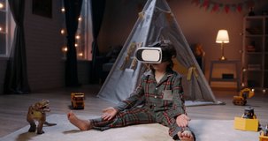 Little boy with curly hair wearing pyjamas is playing video games before bedtime, using virtual reality headset and experiencing new world 4k footage