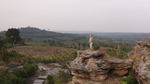 Orbiting shot of a female model with long blonde hair as she's standing on one of the large rocks in the mountain range while she's meditating and doing some yoga exercises alone.