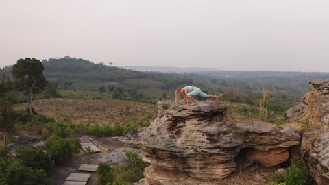 Panning drone shot focused on a large rock by the Cambodian mountains, with a beautiful woman by the rock meditating and exercising yoga on a beautiful sunny day.