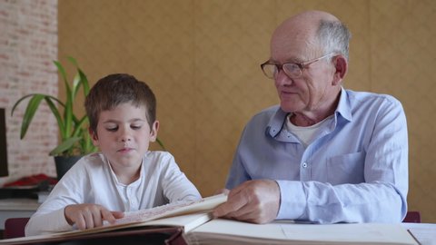 old grandfather in glasses for sight with his sympathetic grandson enjoy memories recall watching a family photo album sitting at a table in a room