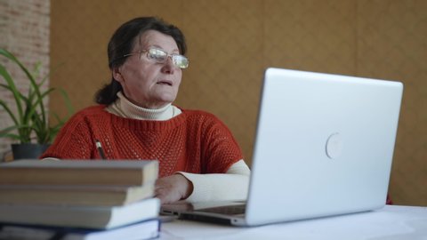 distance studying, aged old woman in glasses for vision uses modern technology is education online via video calling while sitting at table with laptop during training