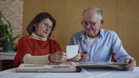 family memories, a cheerful married couple an elderly man and woman enjoy memories flipping through an old family album with photos sitting at a table