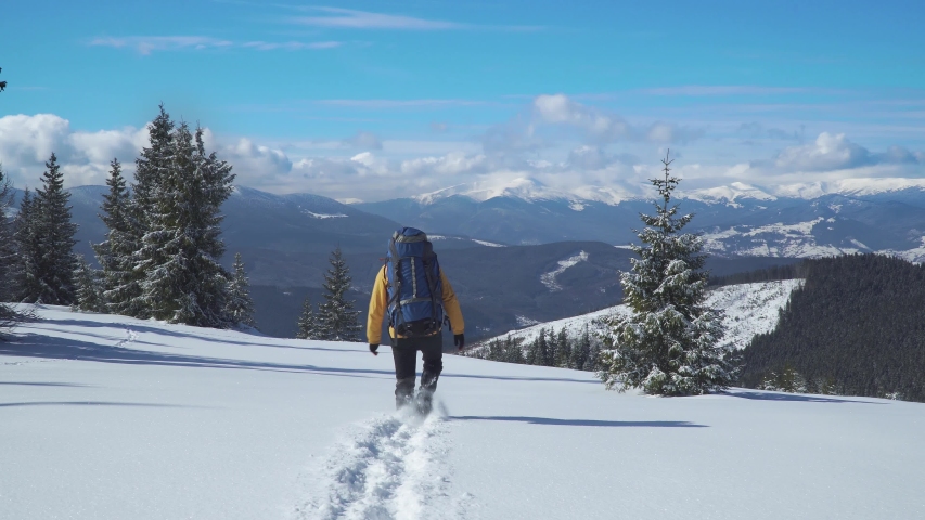 A man with a backpack travels in the mountains in winter. Traveler raising arms victoriously. Carpathian mountain range. The concept of travel and active lifestyle. 4K | Shutterstock HD Video #1054722401